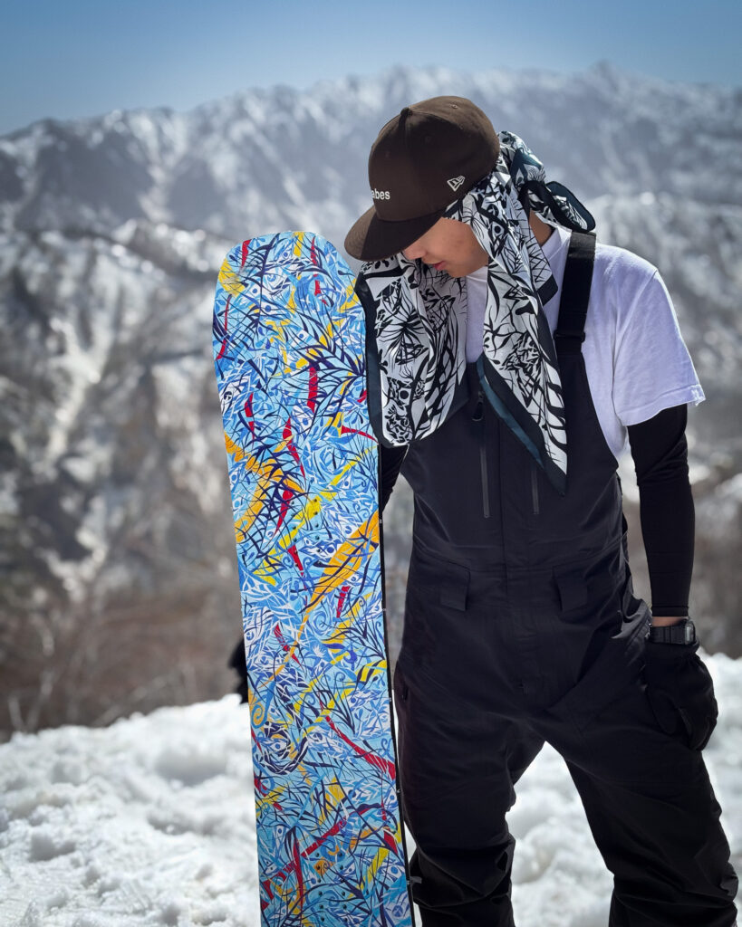 雪山を背景に、アクリル絵具で青を基調に描いたスノーボードアート作品を手にする別府倫太郎