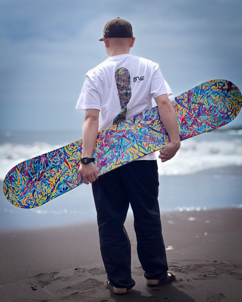 黄色い手描きスノーボードのグラフィックがバックプリントされたTシャツを持っている別府倫太郎