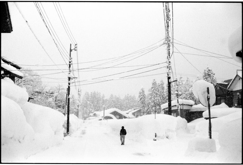 Oratnir(オラトニール）・別府倫太郎による雪国の雪の風景を撮ったモノクロフィルム写真作品。雪の積もった道路の真ん中に祖父が立っているところ。撮影場所:新潟県十日町市 使用カメラ：[Nikon New FM2]、フィルム：[ ILFORD HP5 PLUS 400]